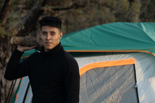 Young man carrying firewood log for a campfire, camping in the forest, standing by a green tent with outdoor survival and adventure concept, enjoying nature at a campsite