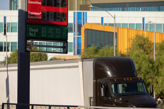 A Gas Station price board displays extremely high gasoline and diesel prices, while a semi tractor trailer freight truck drives behind it.