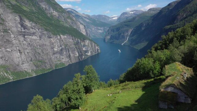 Wide view of a deep fjord surrounded by impressive mountains and a ship, Blomberg G&aring;rd, historic mountain farm on steep mountainside, Geiranger fjord, near Geiranger, M&oslash;re og Romsdal, Norway