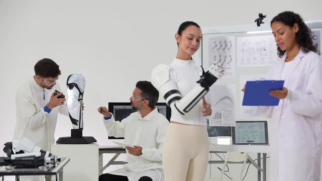Woman with a ponytail flexes a robotic prosthetic hand as a colleague in a lab coat notes results on a clipboard and an engineer examines a robotic leg model