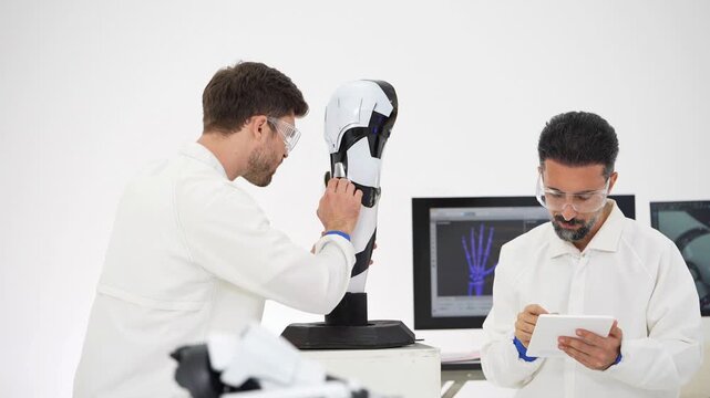 Two engineers in white lab coats and safety glasses study a robotic prosthetic leg model on a stand, one adjusting it while the other uses a tablet beside an X-ray hand monitor
