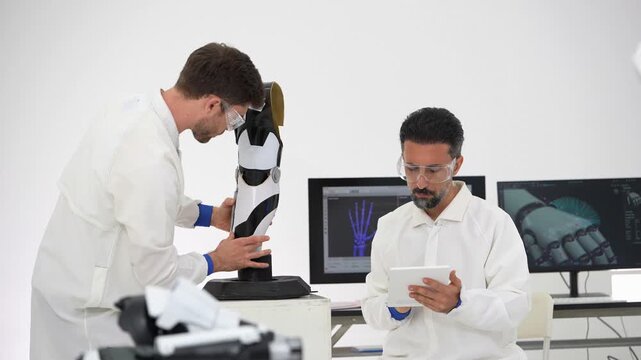 Bearded engineer touches his safety glasses while a colleague leans over a robotic leg model and taps its joints, a hand X-ray monitor glowing behind them