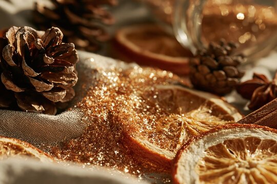 Dried orange slices and pinecones sprinkled with gold glitter. Festive holiday craft supplies are arranged on burlap. Christmas decorations for seasonal projects