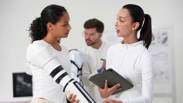 Medium close shot of a curly-haired woman flexing a black robotic prosthetic hand beside a colleague holding a tablet, two engineers working in soft focus behind