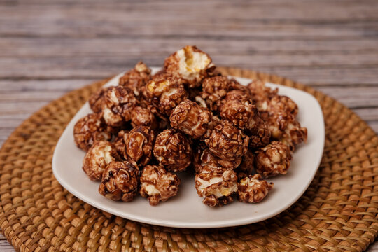 Chocolate popcorn piled high on a white plate. Plate rests on a woven, round mat. Wooden surface beneath adds rustic charm. Sweet, crunchy snack looks deliciously inviting