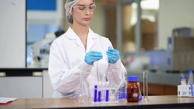 Female technician in a hairnet and white coat carefully pours blue solution between test tubes and a brown reagent bottle on a wooden lab bench, wide shot