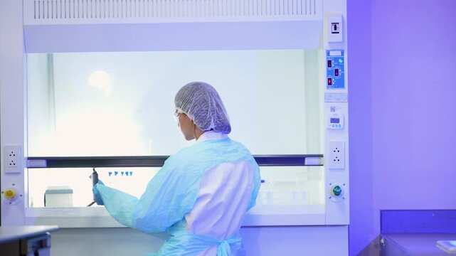 Scientist in a pale blue gown and hairnet stands at a fume hood arranging blue-capped tubes on the counter, seen from behind at a slight angle