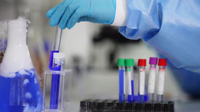 Close-up of test tubes with green, red and purple caps beside a smoking blue flask while blue-gloved hands hold a sample vial in the background