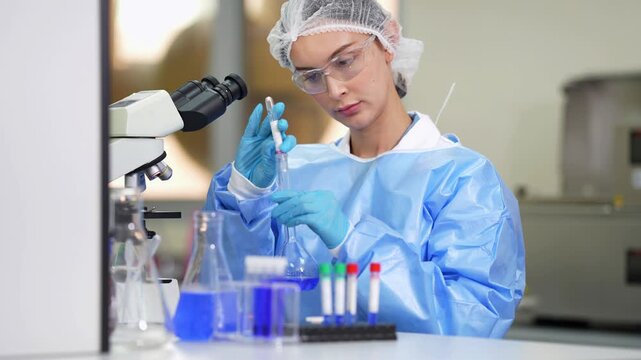 Scientist in blue gown and hairnet inspects a sample tube over a rack of colorful capped tubes, then lifts a flask of blue liquid at a lab bench beside a microscope