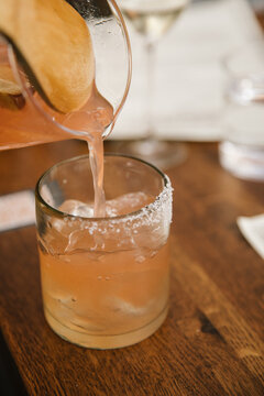 grapefruit margarita being poured from a clear glass pitcher into a clear glass with salt rim and ice on a restaurant table, selective focus