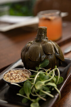 oiled roasted artichoke served with microgreens and dipping sauce with chopped nuts on a restaurant table, selective focus