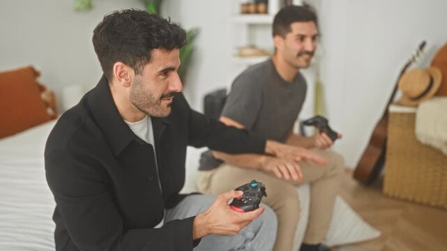 Two men hold game controllers on a bed, leaning forward and smiling while playing in a building; playful competition.
