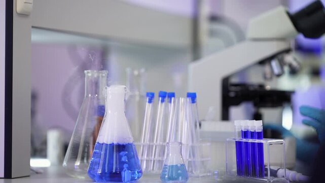 Static close-up of a flask with blue chemical reaction and white vapor beside empty glass tubes and a test tube rack while a gloved hand adjusts a microscope behind