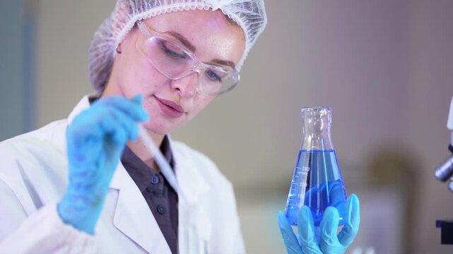 Close-up of a researcher in safety glasses dripping liquid from a pipette into a flask of blue solution, then swirling the flask in blue gloves