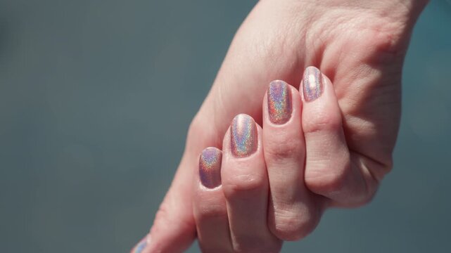 Close up of two hands with iridescent nail polish fingers interlaced in gentle grip. nails display shifting rainbow hues under soft lighting against blurred background. Hands remain steady