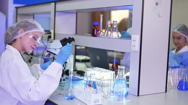 Scientist in a white lab coat pipettes into glassware on a bench beneath shelves of flasks while a colleague in a blue gown works in the background of a modern lab