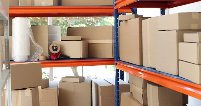 Shelves with many cardboard boxes, bubble wrap and adhesive tape in warehouse. Camera moving left