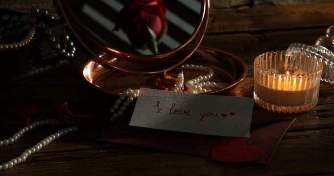 Card with phrase I Love You, envelope, burning candle, accessories and mirror on wooden table, closeup. Changing direction of light