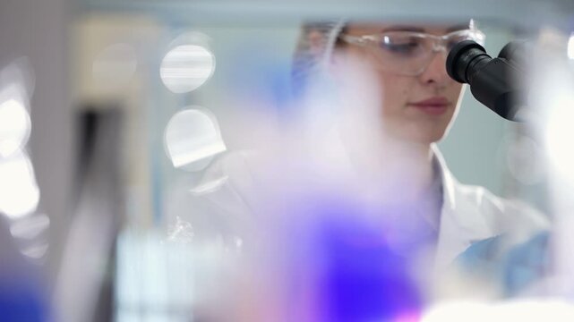 Female scientist peers into a microscope through a soft blurred foreground of glassware and purple liquid, close-up of her face in a laboratory