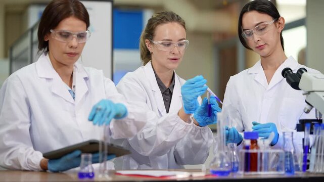 Team of three female scientists handle pipettes, flasks and test tubes at a lab table while one holds a tablet, blue reagents in the foreground, medium shot