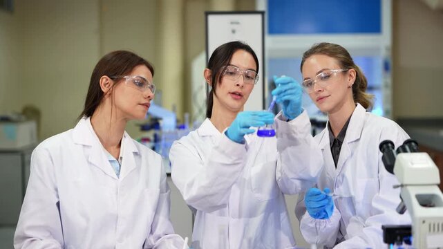 Three female scientists stand in a lab as the middle one mixes blue solution in a beaker with a stirring rod while colleagues look on beside a microscope