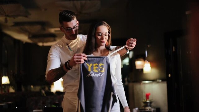 Man stands behind woman and guides dark jacket onto her shoulders with both hands while she faces forward with calm confident expression, visible action focuses on jacket placement and woman holding