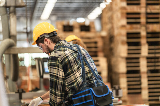 Bearded woodworker in a yellow hard hat and plaid shirt pauses his cutting work to check the line with towering stacks of wooden pallets behind him in the era of AI and robotic automation