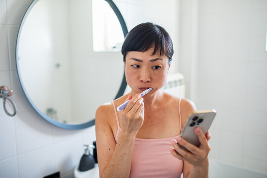 Woman brushing teeth while looking at smartphone in bathroom