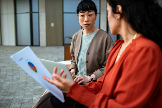 Two coworkers discuss a pie chart outdoors with a laptop
