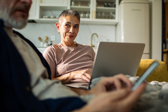 Senior woman using laptop at home beside man on phone