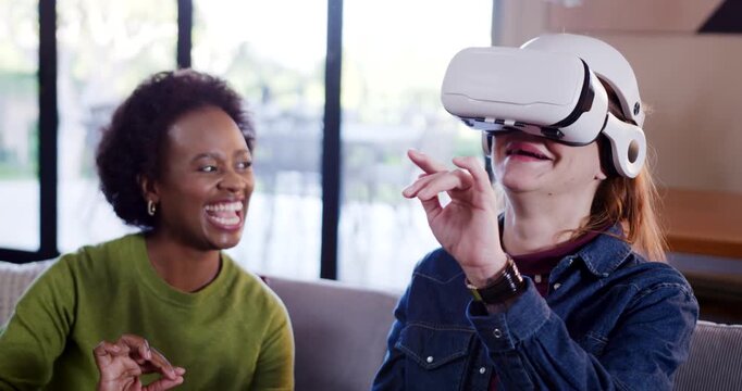 Diverse female friends sitting on sofa, right woman putting on VR headset, exploring virtual menu