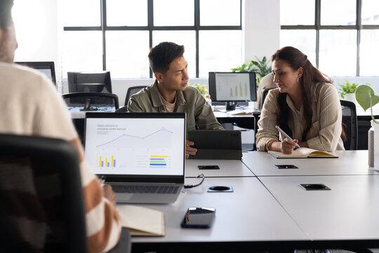 Diverse coworkers collaborating while reviewing laptop charts and tablet at open-plan office table