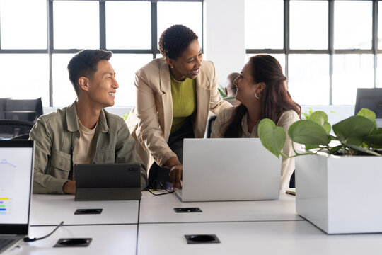 Diverse coworkers collaborating at shared desk in open plan office using laptops and tablet