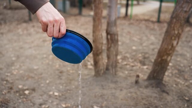 Hand holds blue collapsible container pouring water onto ground in outdoor setting with trees and dirt visible. action shows liquid flowing steadily from cup as it is tilted demonstrating