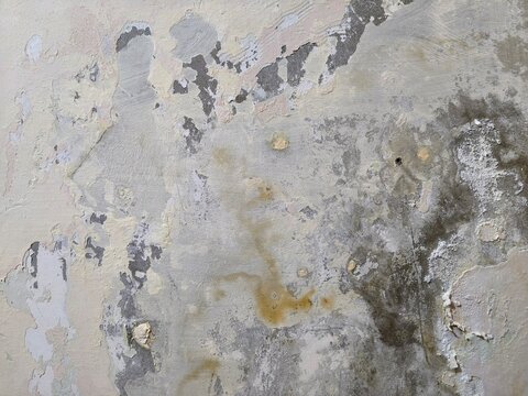 A close-up of a weathered concrete wall with peeling and chipped paint. The surface shows layers of faded beige, gray, and white paint flaking off to reveal the rough concrete underneath.