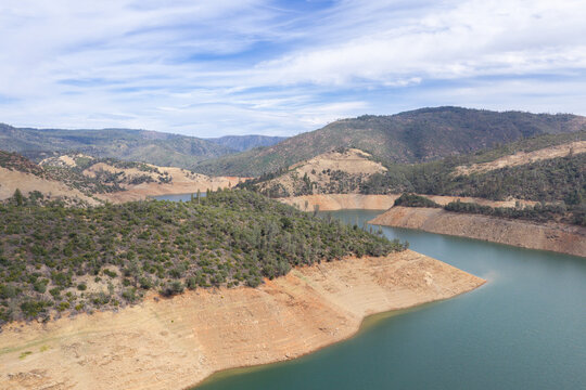 Lake Oroville experiencing significant drought conditions with low water levels exposing dry, cracked earth on the shoreline, surrounded by hills and a blue sky