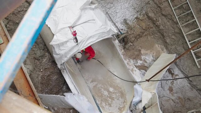 Worker cuts an opening in a fiberglass-reinforced cured-in-place pipe liner during sewer rehabilitation, wearing protective clothing and safety equipment inside an excavation.