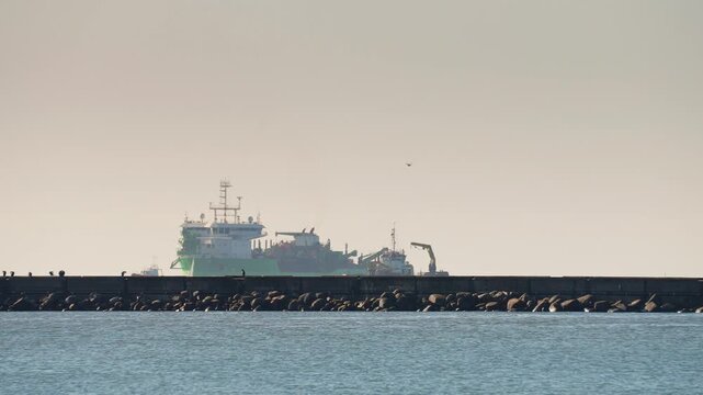 Large construction vessels and workboats operating behind a breakwater during major marine infrastructure works, with cranes, barges and harbour activity in hazy coastal light.