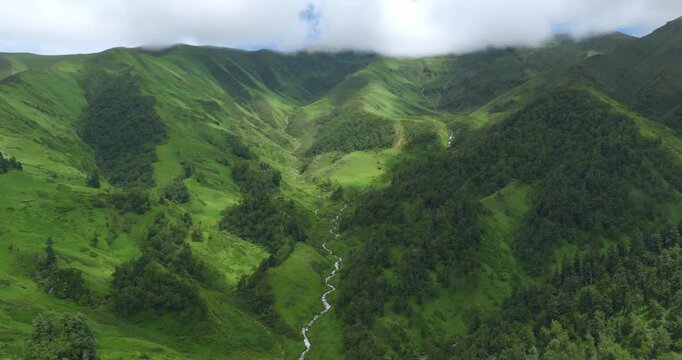 Badimalika, Bajura. Nepal, drone hyperlapse shot of a winding river stream flowing through lush alpine slopes and pine forests as mesmerizing monsoon clouds roll over green mountain ridges.