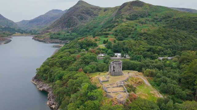 Ancient stone fortress standing above llyn lake waters, camera pulling back elevated above wooded hillsides and mountain backdrop