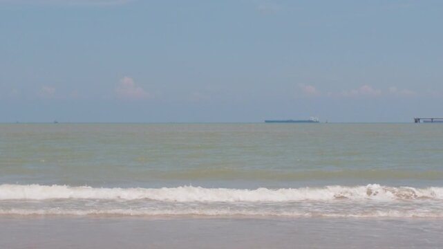 Static 4K slow motion shot of gentle waves washing onto the sandy shore at Pantai Puteri in Melaka, with a distant container ship on the horizon.