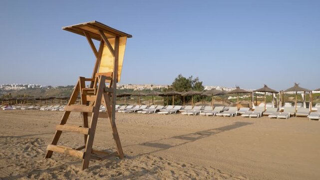 Wooden lifeguard tower stands empty on a sunny beach as human shadows walk in the foreground. Background shows sunbeds and parasols by the sea. Calm summer morning and relaxing coastal mood.