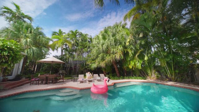 Beautiful girl relaxing by the pool on a sun lounger in a tropical backyard. Pink flamingo float on turquoise water. Tall palm trees, patio umbrella and dining table under blue sky on a sunny day.