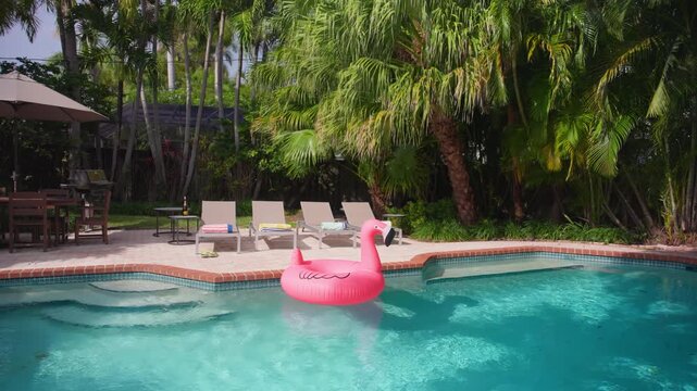 Pink flamingo float drifts in a turquoise pool. Relaxing by the pool in a tropical backyard with four sun loungers, striped towels, flip flops, palm trees and a patio umbrella. Sunny summer day.