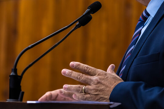 Close-up of a businessman or politician wearing a green suit and watch gesturing during a speech with microphones at a podium. 