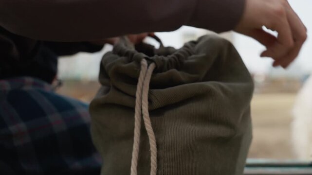 Person hands adjust drawstring on green fabric bag revealing texture details. background is softly blurred with hints of outdoor setting and another person in plaid clothing nearby. Soft