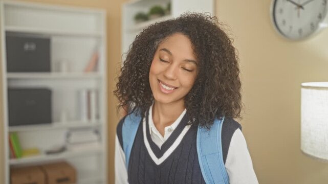 Woman student in school uniform with blue backpack smiling and tilting head by bookshelf and wall clock inside a building study area; cheerful curiosity.