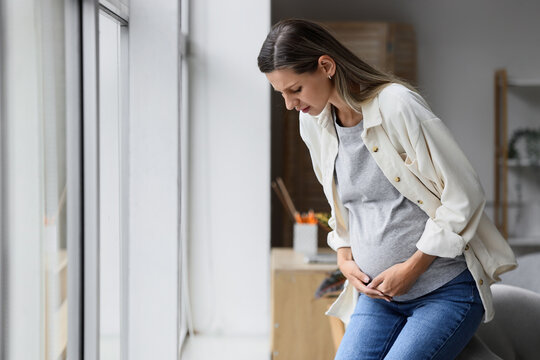 Pregnancy pain and discomfort, pregnant woman feeling abdominal cramps or contractions while standing at home near a window