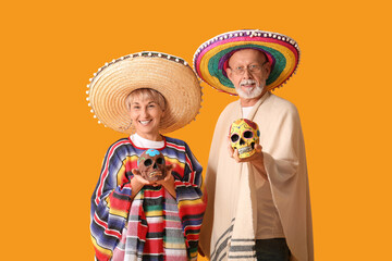 Obraz premium Dia de los muertos celebration, happy senior couple wearing traditional mexican sombreros and ponchos holding decorated sugar skulls on orange background