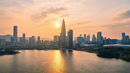 Obraz premium A modern urban skyline at sunset, featuring tall skyscrapers and their reflection on calm waterfront under warm orange sky.